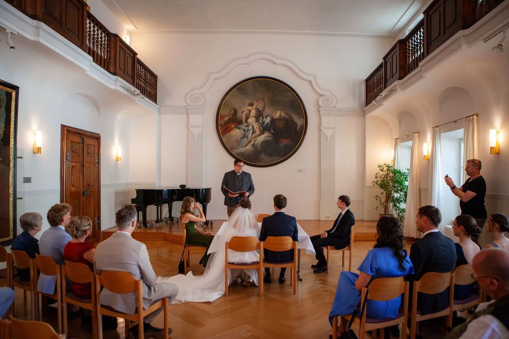 Small intimate wedding ceremony in elegant hall with large painting, guests seated in wooden chairs.