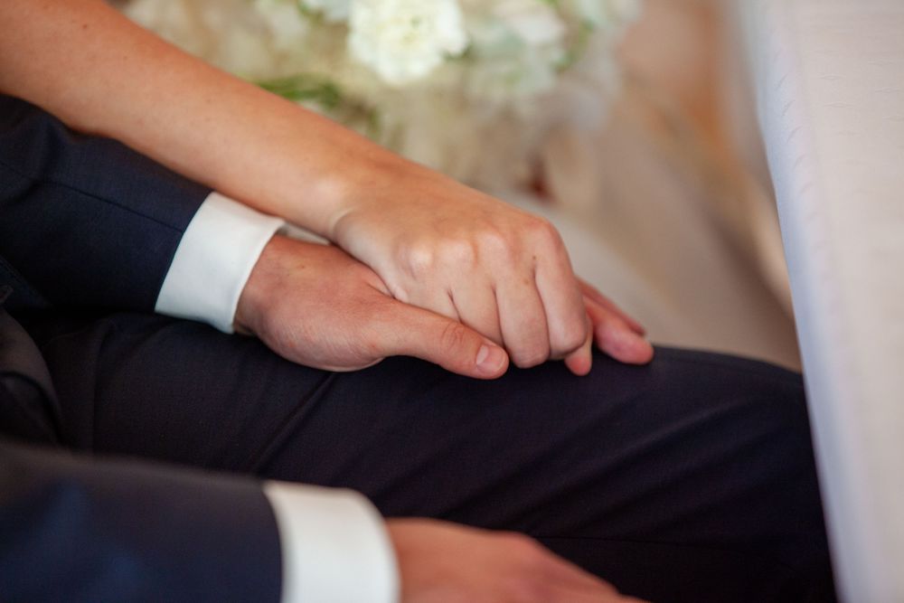 Close-up of bride and groom holding hands during wedding ceremony, groom wearing dark suit.