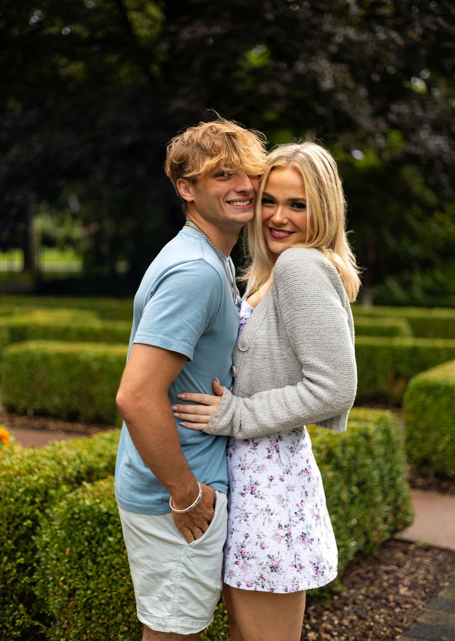 Smiling young couple embracing in a formal garden with manicured hedges and trees.
