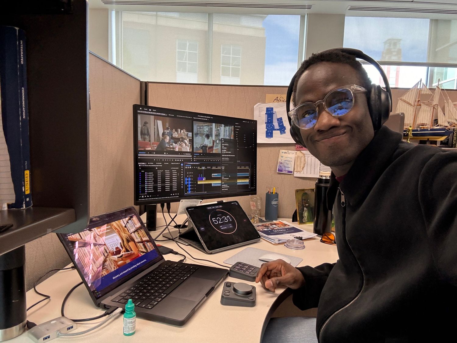 A smiling person wearing headphones sits at a multi-monitor video editing workstation in a modern office cubicle.