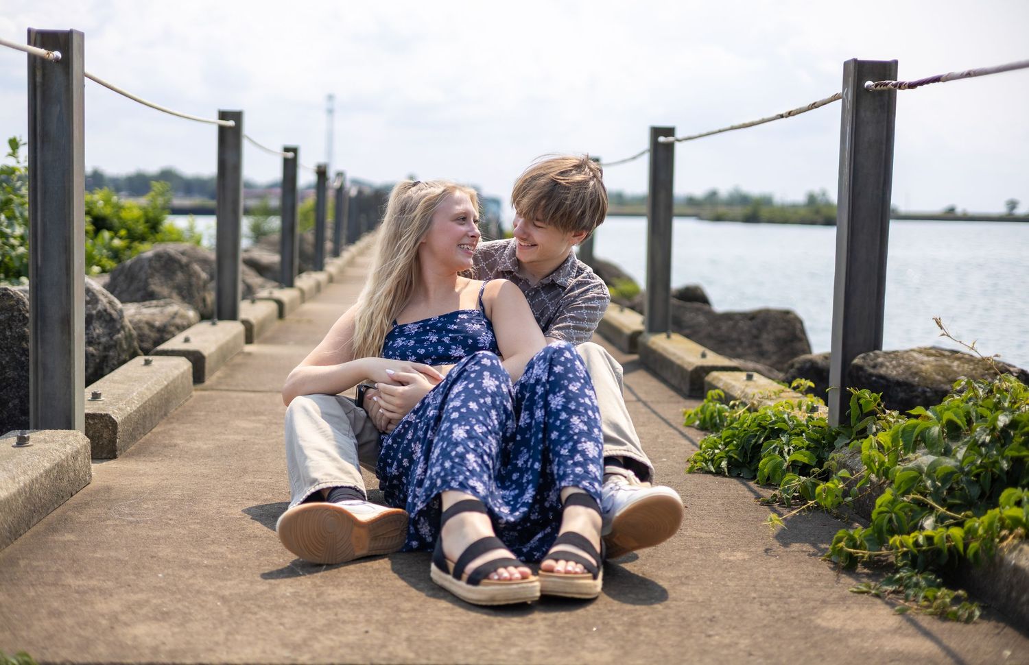 Couple sitting together on waterfront pathway, sharing a romantic moment by the water on a sunny day.