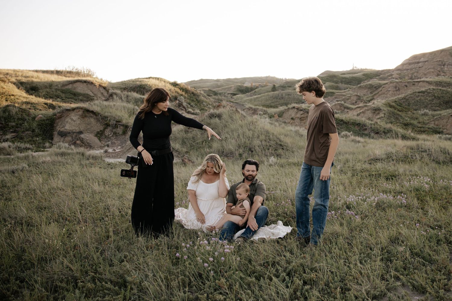 Two photographers capturing a woman in white dress with children near a farm fence at golden hour.