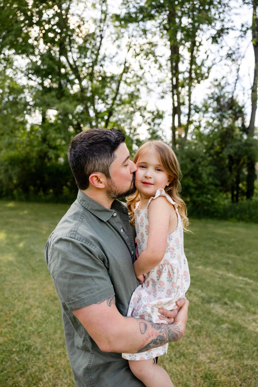 A father kisses his young daughter on the cheek while holding her in a lush green park setting.