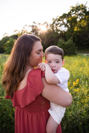 A mother in a red dress holds her young son in a sunlit green field at golden hour.