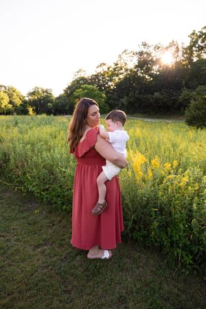 Woman in red dress holding a baby in a sunny green field with yellow wildflowers at golden hour.