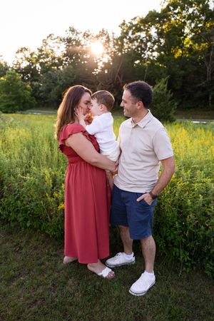 Family portrait at golden hour, mother in red dress and father in beige polo holding toddler in white outfit outdoors.