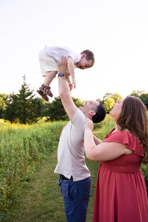 A joyful father lifts his toddler high in the air outdoors while mother watches and smiles in a green field.
