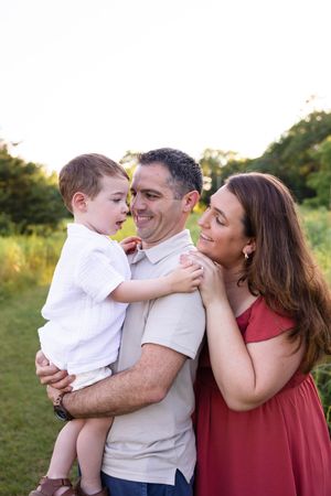 A happy family of three poses outdoors, with father holding young son while mother smiles beside them in a green field.