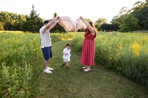Family of three playing with a blanket in a sunny green field, parents lifting fabric over toddler outdoors.