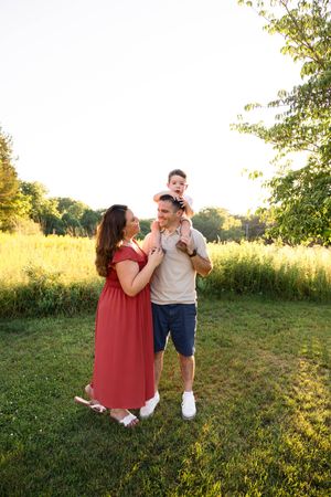 Family of three enjoying golden hour outdoors, mom in red dress, dad carrying toddler on shoulders in green park.