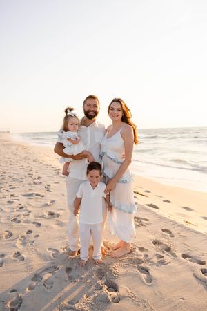 Happy family of four in white outfits posing on a sandy beach at sunset with ocean waves behind them.