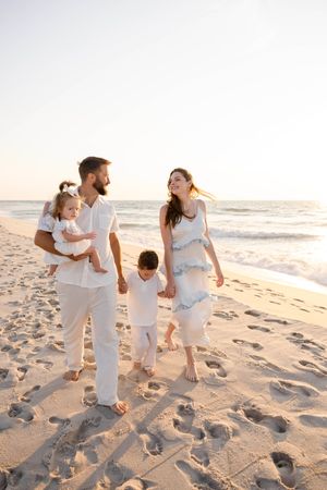 Happy family of four walking on a sunny beach, parents holding children's hands, dressed in white outfits.