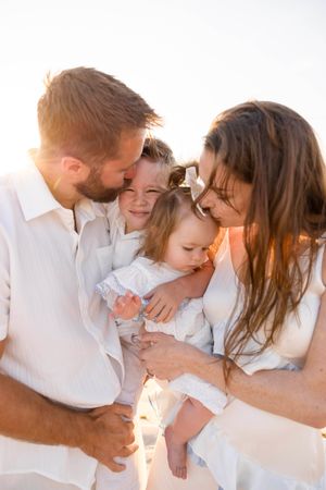 Family of four in white outfits sharing a tender moment on a sunlit beach at golden hour.