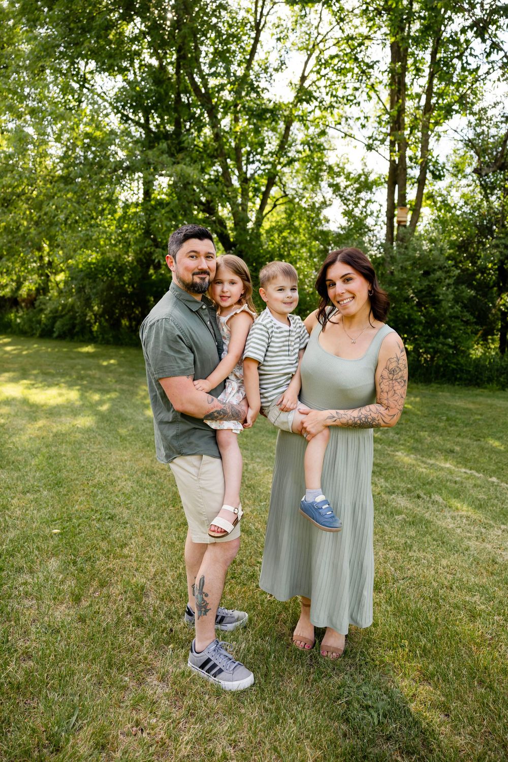 A happy family of four poses outdoors on green grass, parents holding two young children, surrounded by lush trees.