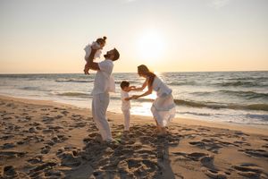 Family in white outfits playing on a sunny beach at sunset, with waves and golden sand in the background.