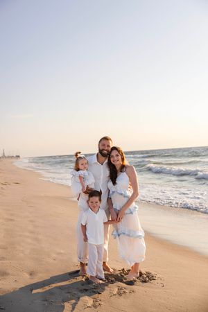 Family of four in white outfits posing on a sandy beach at sunset with ocean waves in the background.