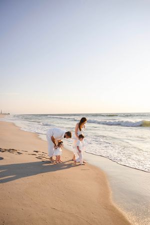 Family in white outfits enjoying a beach walk at sunset near ocean waves on sandy shore.