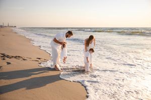 Family in white outfits playing in ocean waves on a sandy beach at sunset.