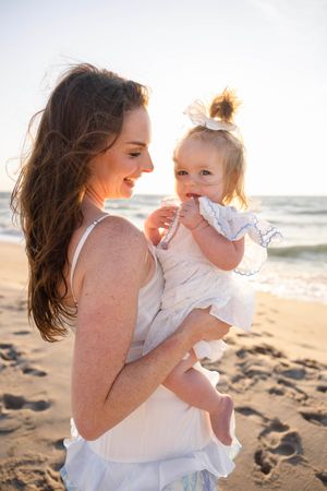 A mother in a white dress holds her baby girl on a sunny beach, both dressed in white with ocean waves behind them.