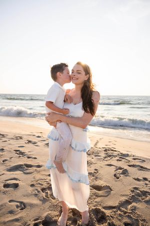 A mother in a white dress holds her young son on a sunny beach as he kisses her cheek near ocean waves.