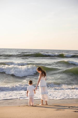 Mother and young child in white outfits holding hands on sandy beach with ocean waves at sunset.
