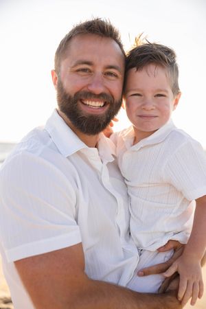 Smiling bearded father holding young son on beach, both wearing white shirts in warm sunlight.