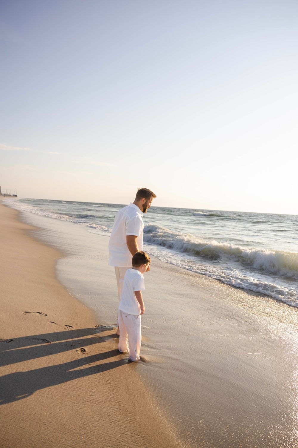 Father and young son in matching white outfits standing together at the ocean's edge on a sandy beach at sunset.
