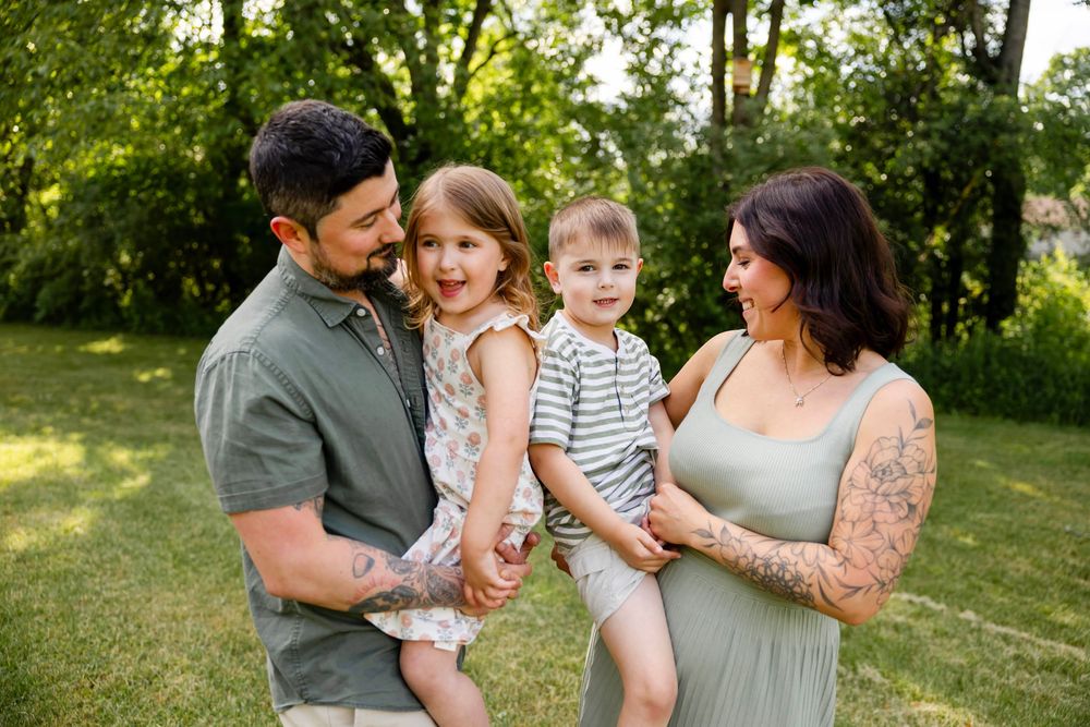 Happy family of four posing outdoors in a lush green park, parents holding young children, mother visibly pregnant.
