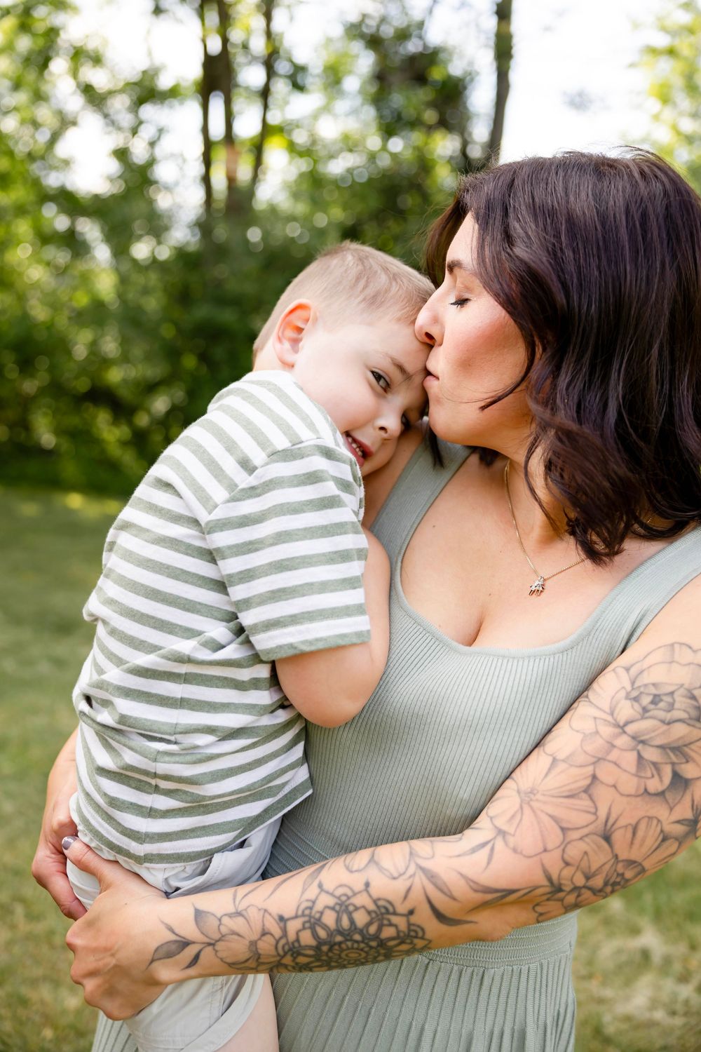 A tattooed mother kisses her young toddler son while holding him outdoors in a lush green park setting.