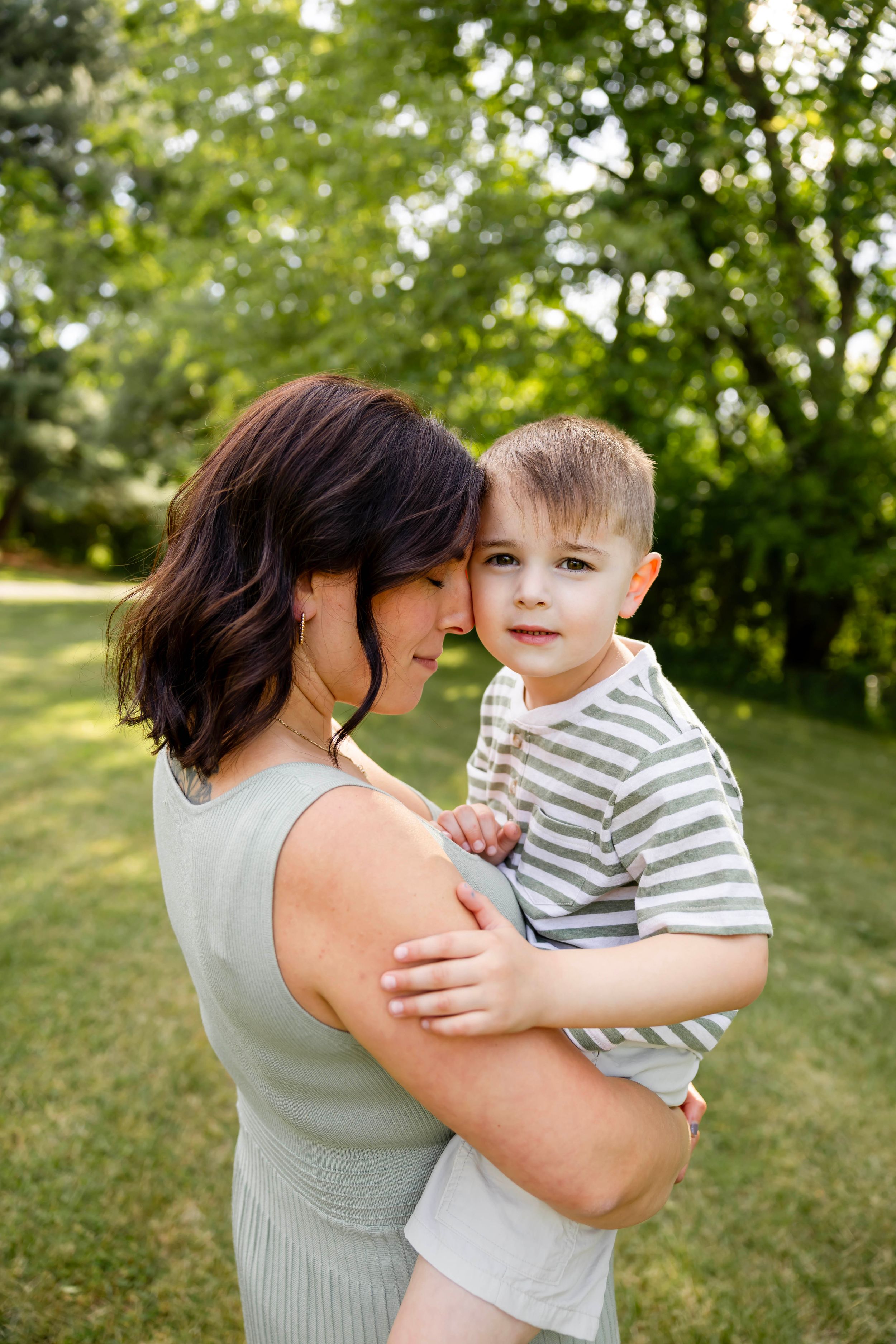 A mother holds her young son in a sunny green park, both smiling warmly in a tender outdoor family portrait.