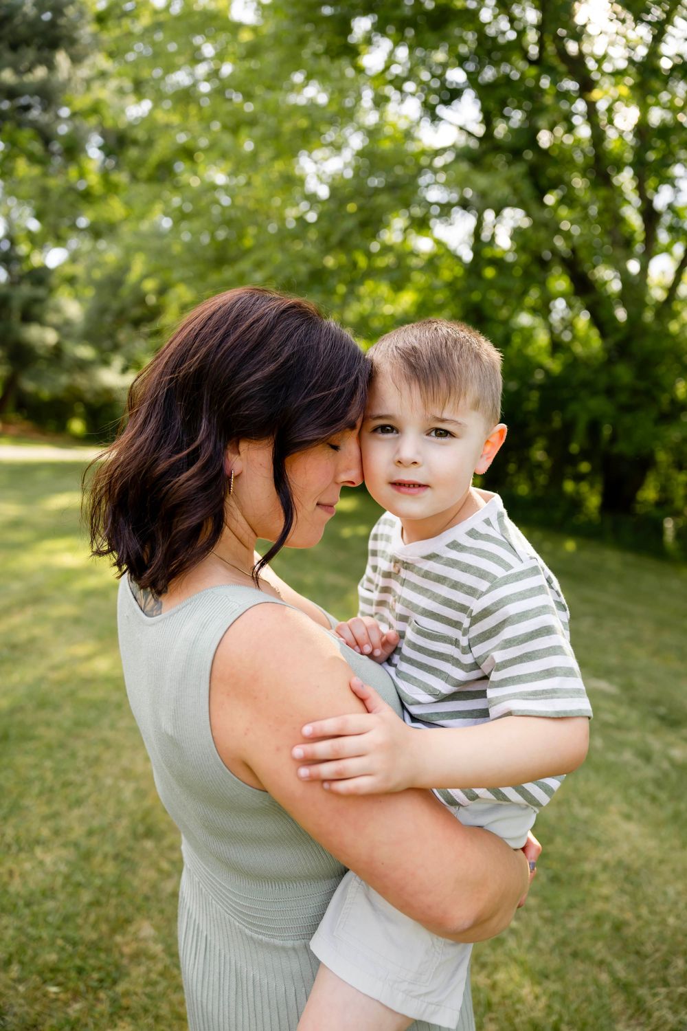 A mother holds her young son in a sunny green park, both smiling warmly in a tender outdoor family portrait.
