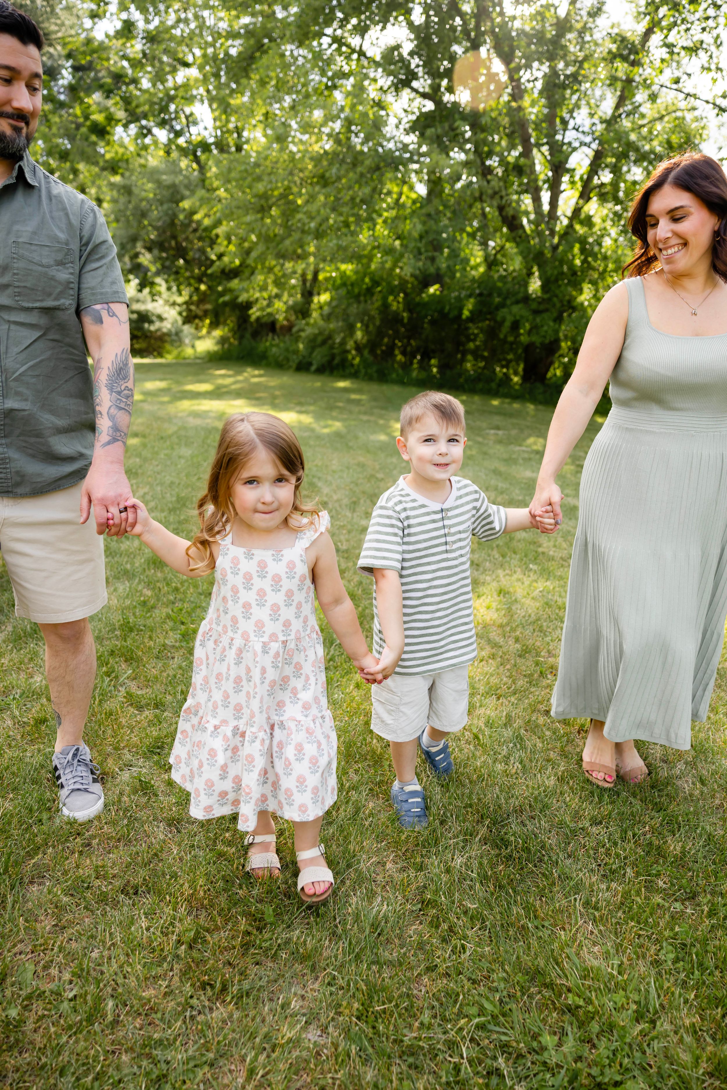 Happy family of four holding hands and walking together on green grass in a sunny backyard park setting.
