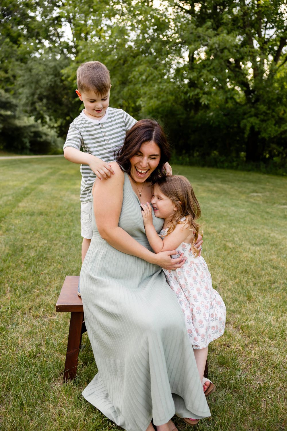 A laughing mother in a mint dress sits on a bench outdoors, hugged by her two young children in a lush green yard.