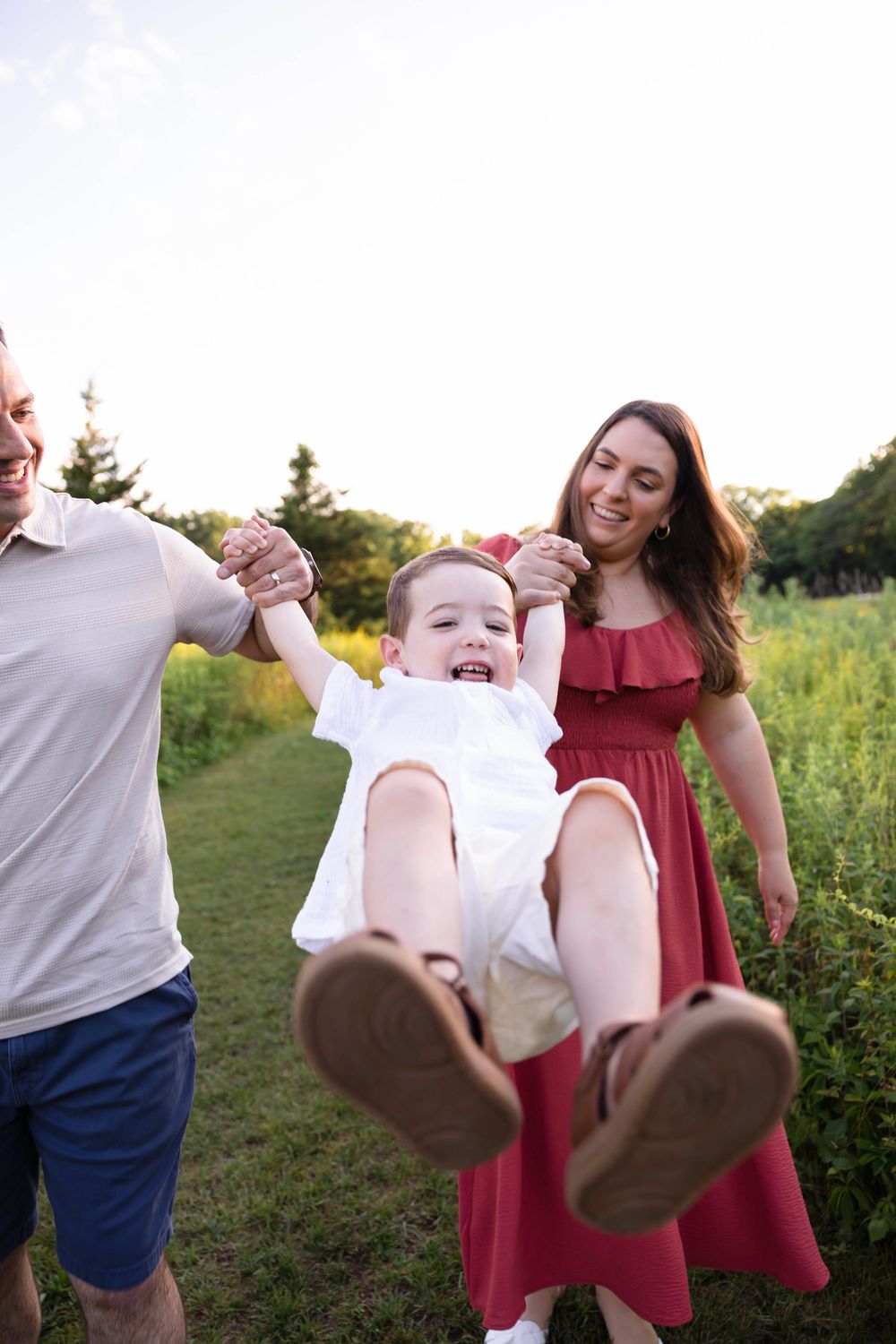 Happy family swinging toddler outdoors in a sunny green field, parents smiling as child laughs with joy.