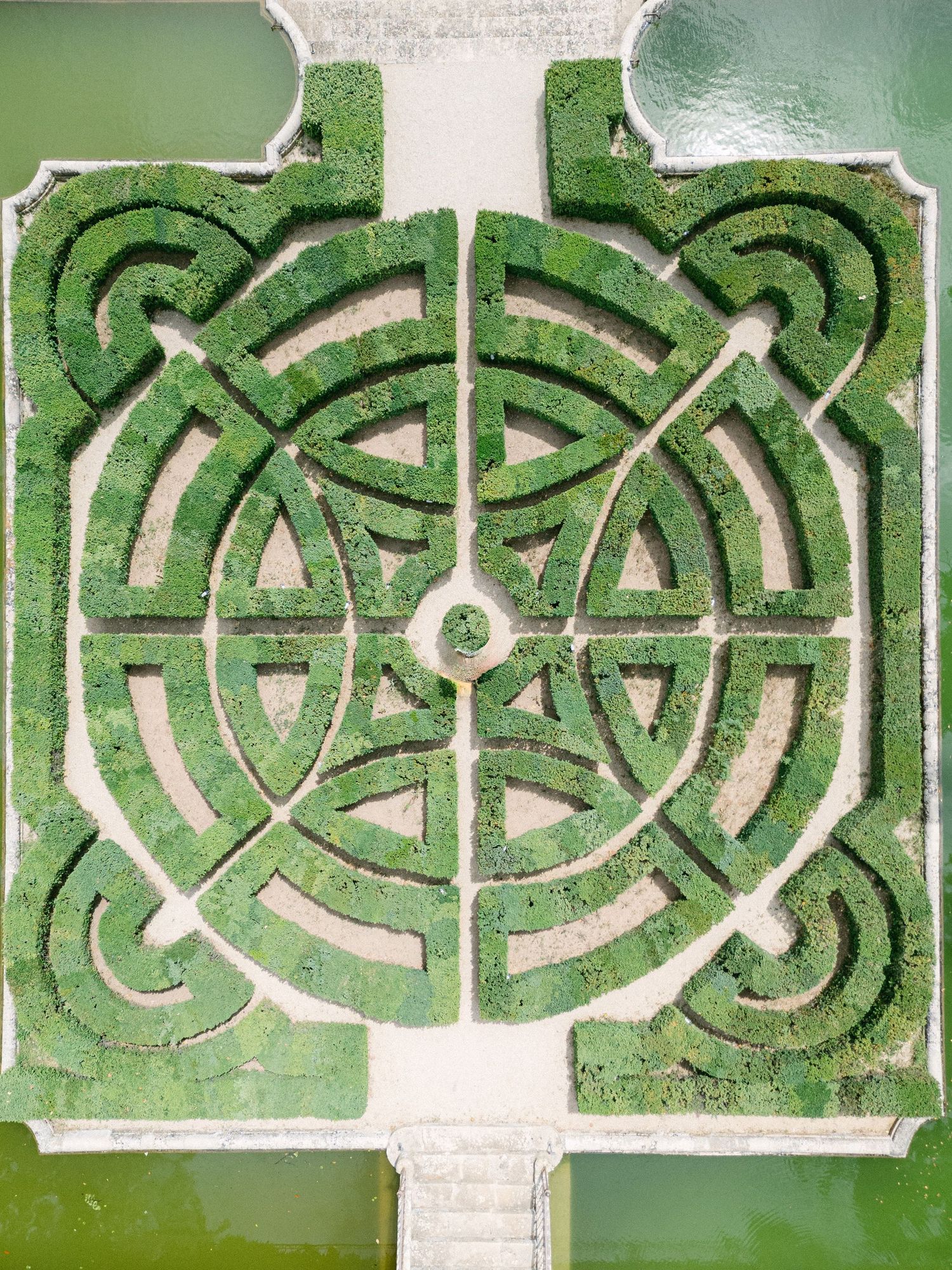 Aerial view of an intricate green hedge maze with geometric patterns, surrounded by water features.