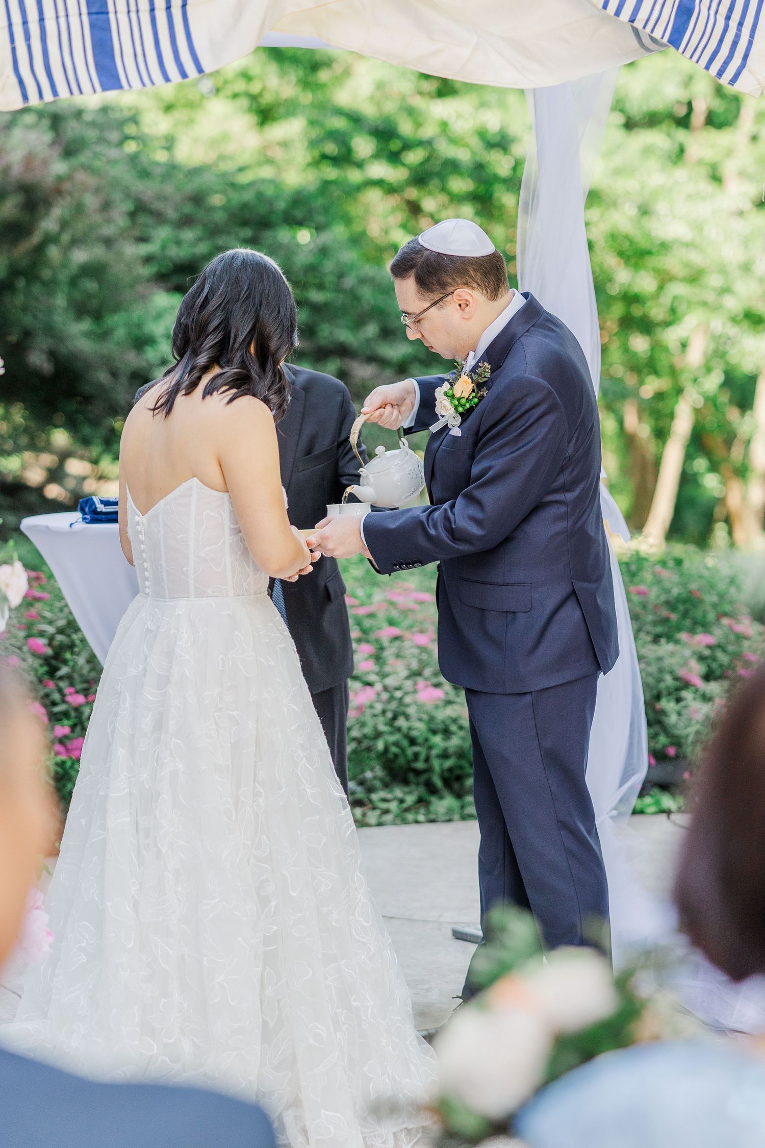 Bride and groom exchange rings under a chuppah during an outdoor Jewish wedding ceremony surrounded by lush greenery.