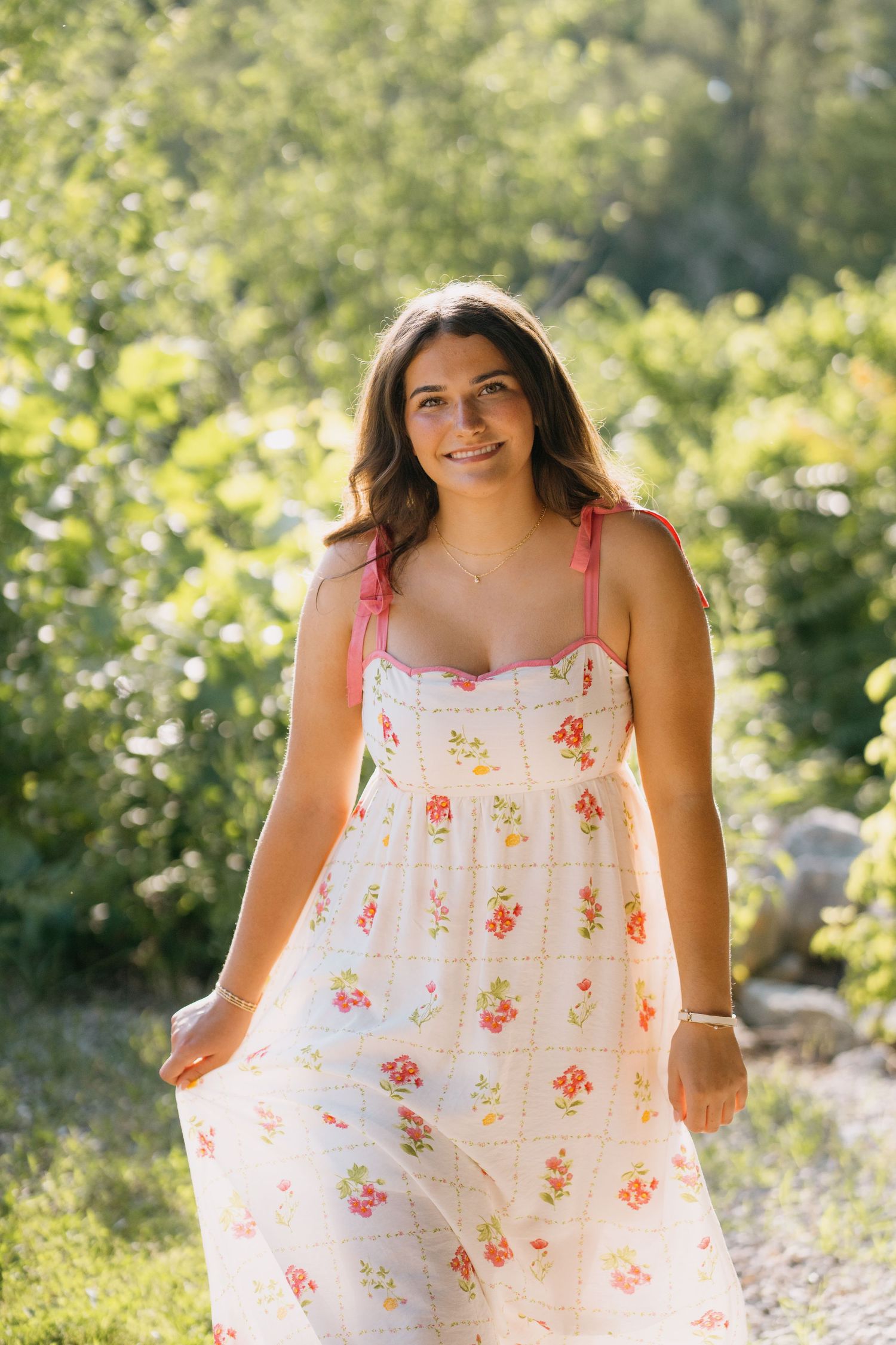Young woman smiling in a floral sundress outdoors, surrounded by lush green trees in warm sunlight.