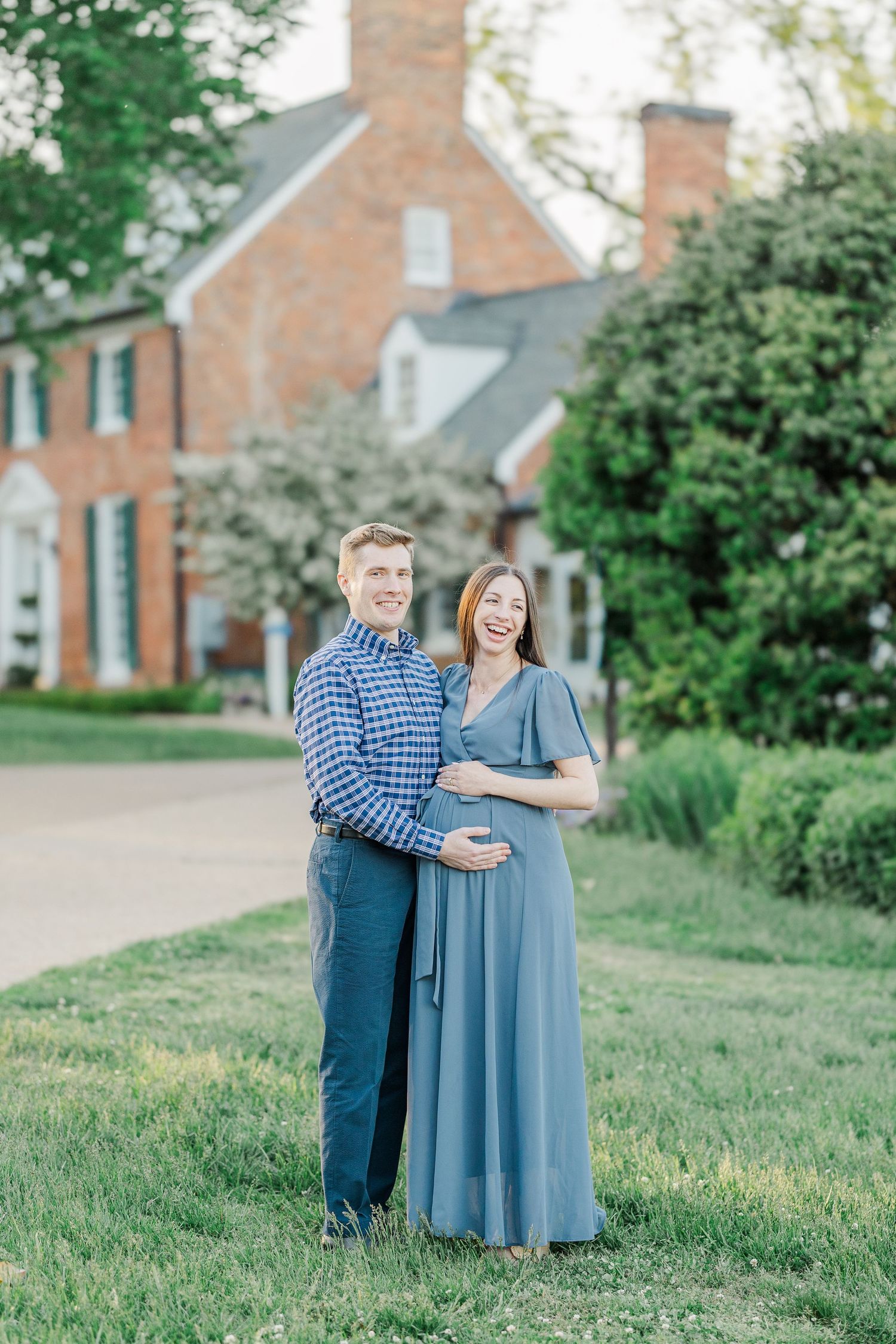 A smiling couple poses outdoors near a brick estate, the woman in a blue maternity gown cradling her bump.