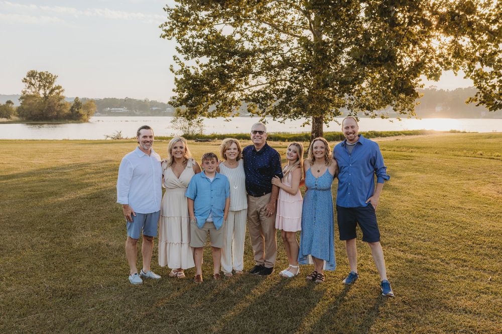 Family of eight posing together on green lawn near lake under large tree at golden hour sunset.
