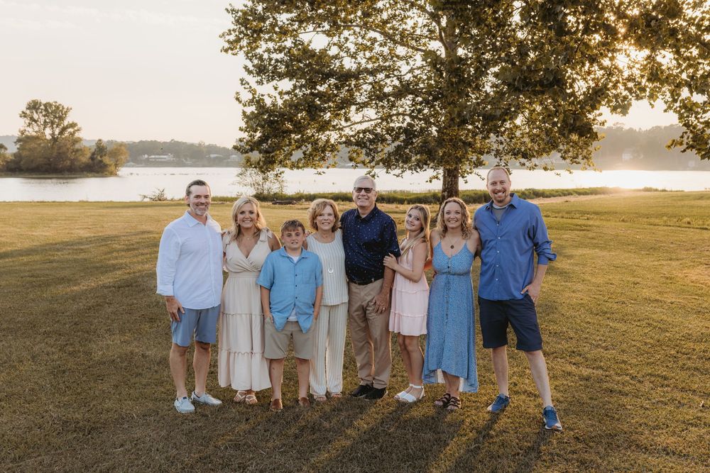 Family of eight posing outdoors near a lake at golden hour on a grassy lawn with a large tree backdrop.