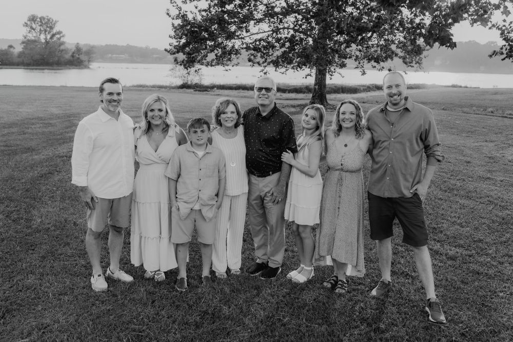 Black and white family portrait of eight people smiling outdoors near a tree and lake.