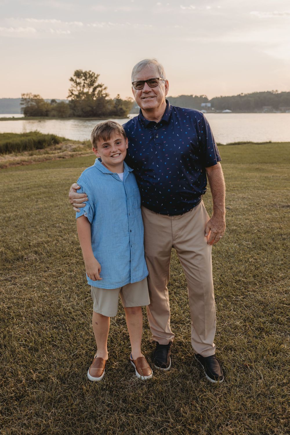 A grandfather and young grandson pose together smiling on a grassy lawn near a lake at sunset.