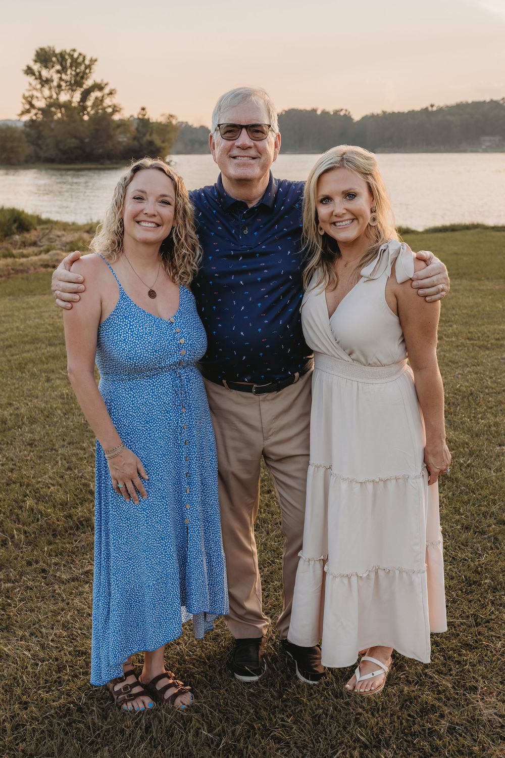A man and two women smiling together outdoors near a lake at sunset, wearing summer attire on a green lawn.
