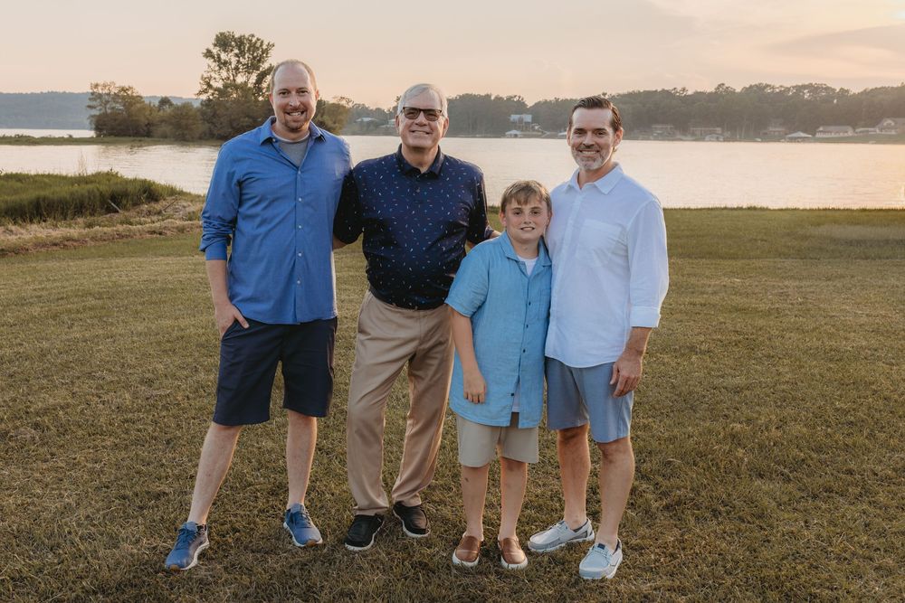 Four males pose smiling outdoors near a lake at sunset on a green lawn, wearing casual blue shirts and shorts.