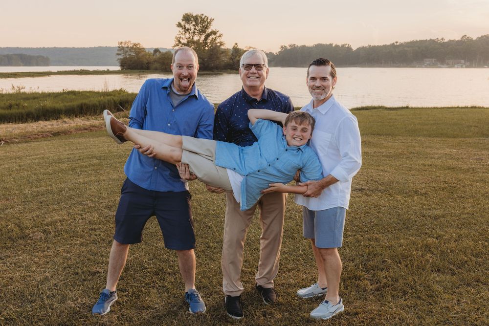 Four men playfully pose outdoors near a lake at sunset, with three holding one man horizontally while smiling.