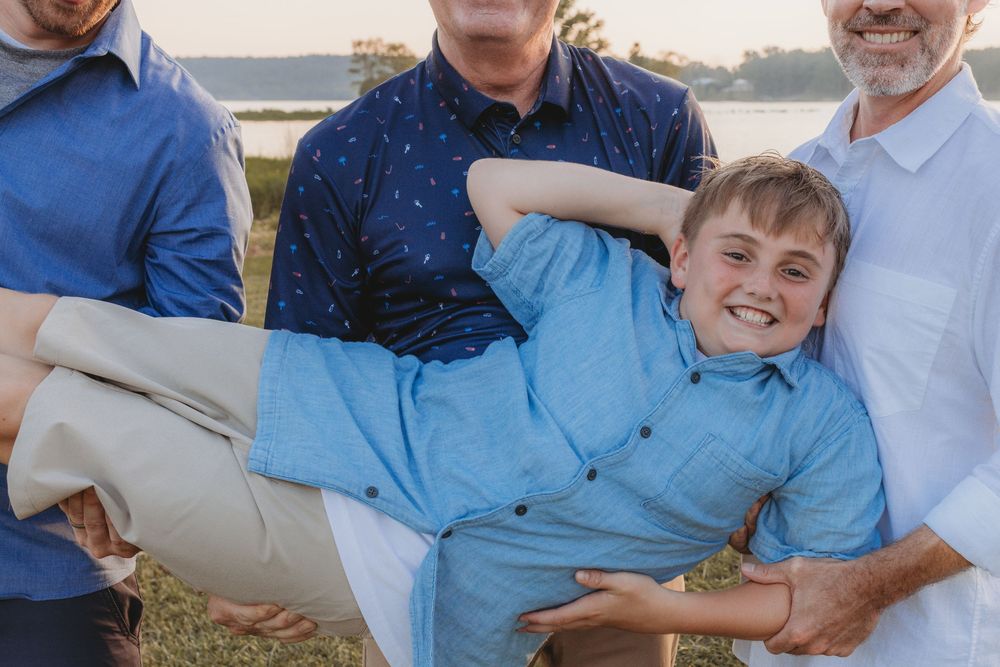 Two men carrying a smiling boy in a blue shirt outdoors near a lake during a family photo session.