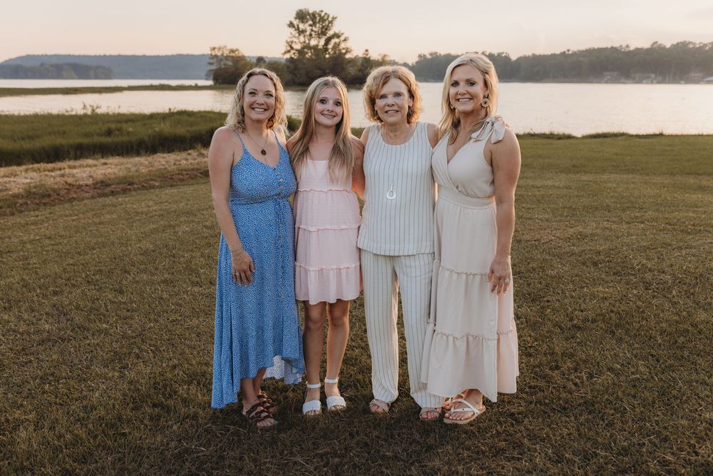 Four women smiling together outdoors near a lake at sunset on a green lawn.