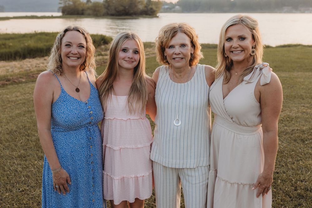 Four women smiling together outdoors near a lake at golden hour, wearing summer dresses in blue, pink, white, and cream.