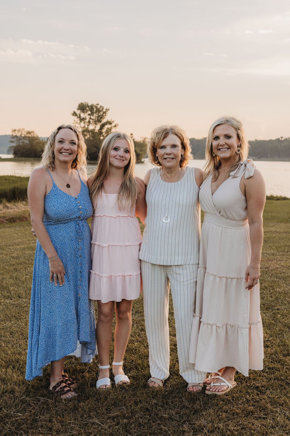 Four women smiling outdoors near a lake at sunset, wearing elegant summer dresses.