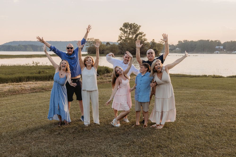A joyful group of eight people celebrating outdoors near a lake at sunset, arms raised in excitement on a grassy lawn.